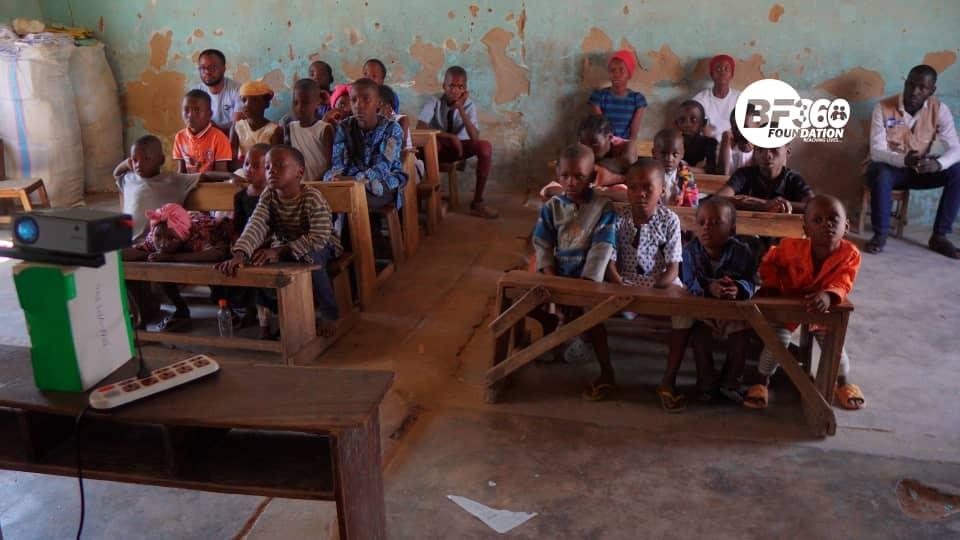 A Diyaa projector on a wooden desk facing a classroom of children seated on benches in a rural school